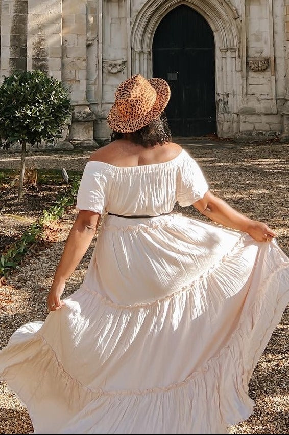 woman wearing a flowy off-the-shoulder cream maxi dress with a tiered hem and a leopard print hat