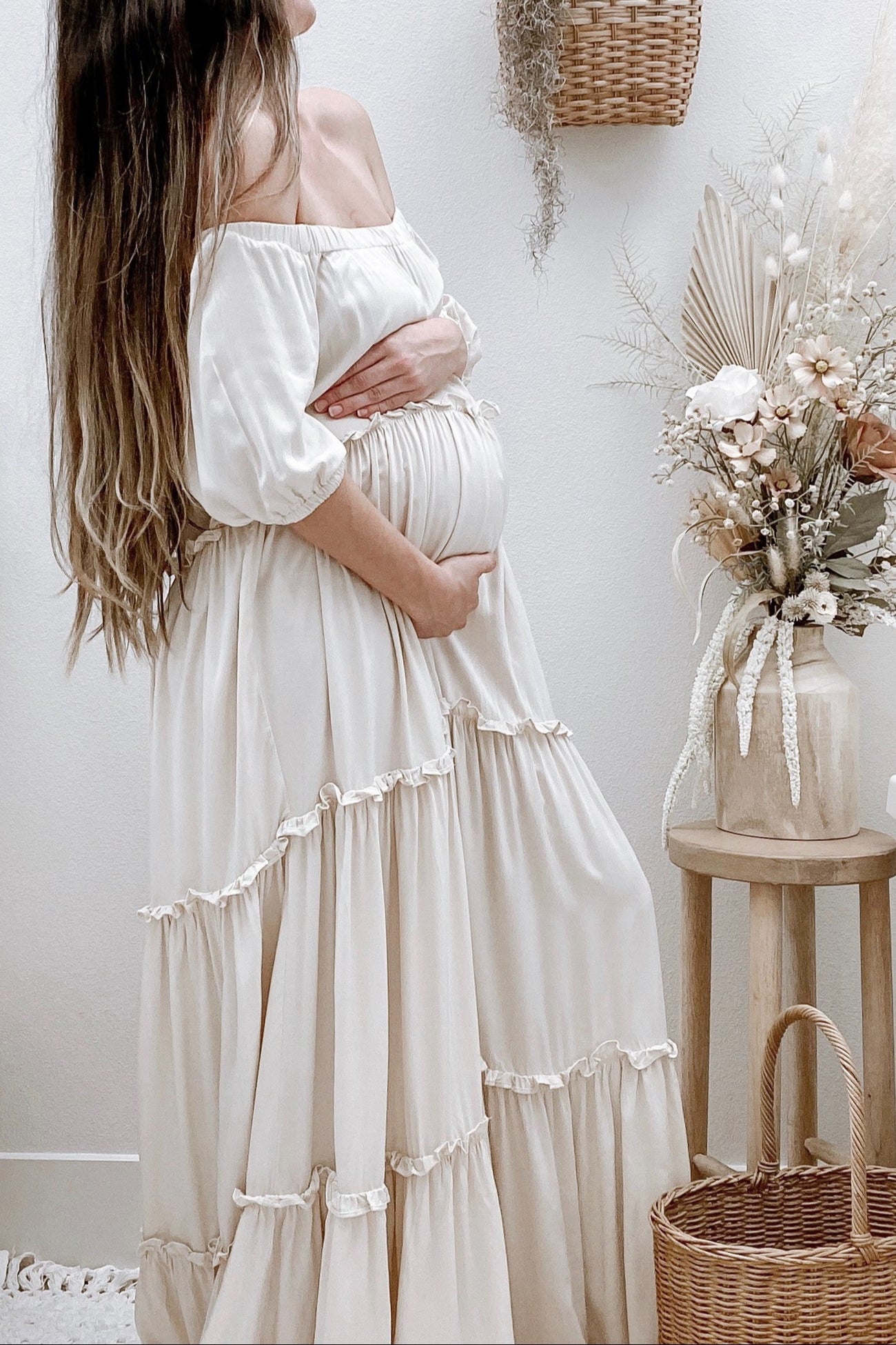 woman wearing a cream off-the-shoulder maternity dress with ruffle details and holding her baby bump