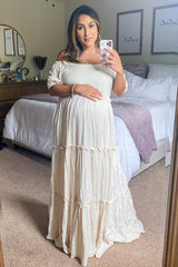 woman wearing a cream off-the-shoulder maternity maxi dress and holding her baby bump