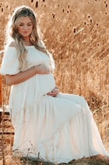 woman wearing a white off-the-shoulder maternity dress sitting in a field, holding her baby bump