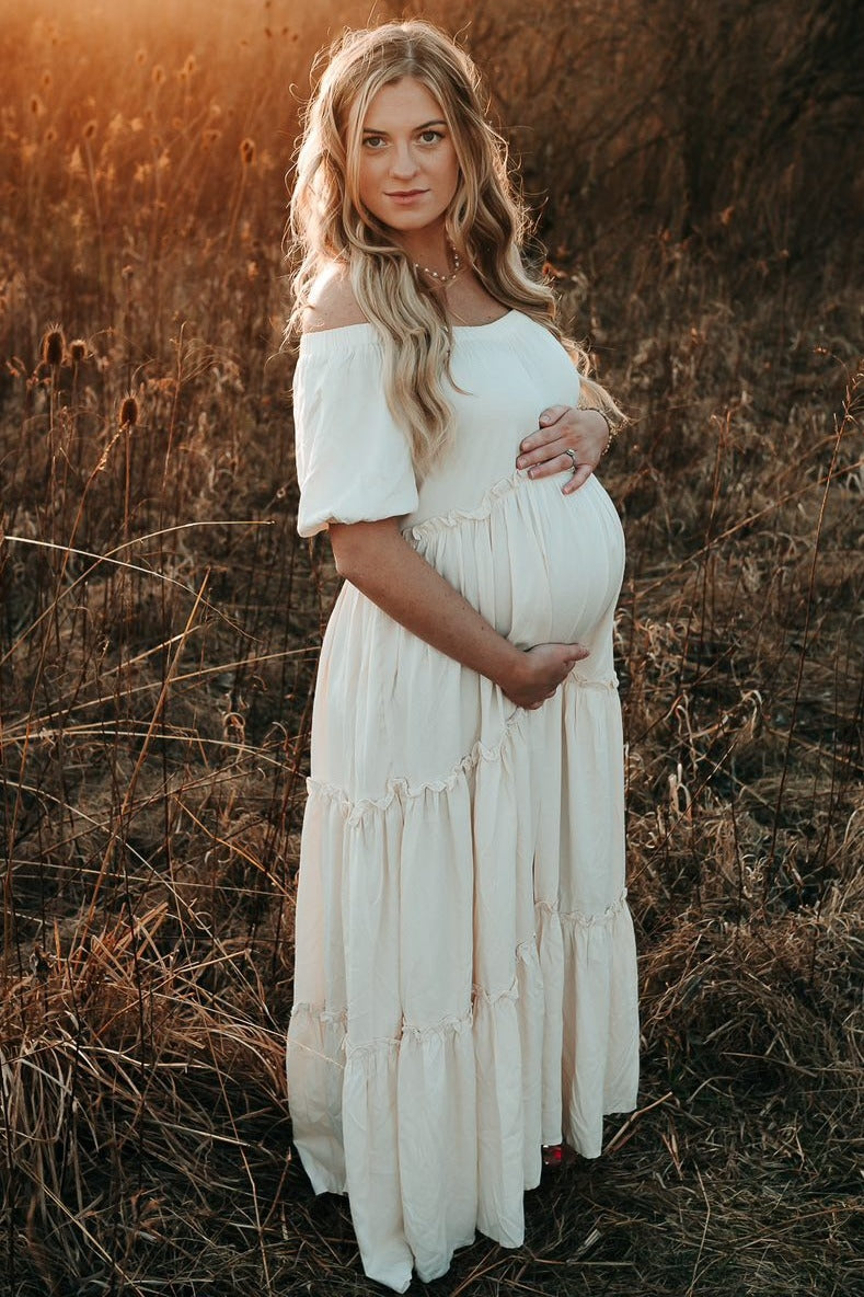 woman wearing a cream off-the-shoulder maternity dress with tiered details and holding her baby bump outdoors