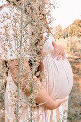 woman wearing a light pink maternity dress and holding her baby bump among plants