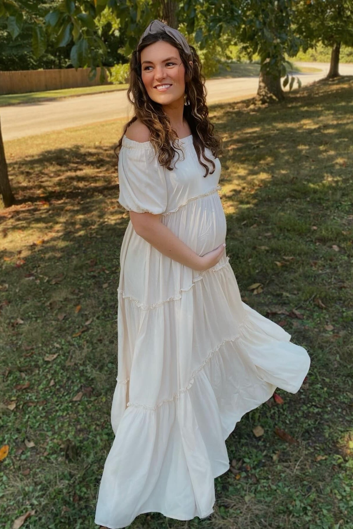 woman wearing a flowy white off-shoulder maternity dress, holding her baby bump in a green outdoor setting
