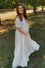 woman wearing a flowy white off-shoulder maternity dress, holding her baby bump in a green outdoor setting