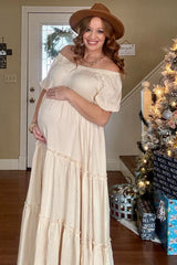 woman wearing a cream off-the-shoulder maternity maxi dress and showcasing her baby bump, accessorized with a hat