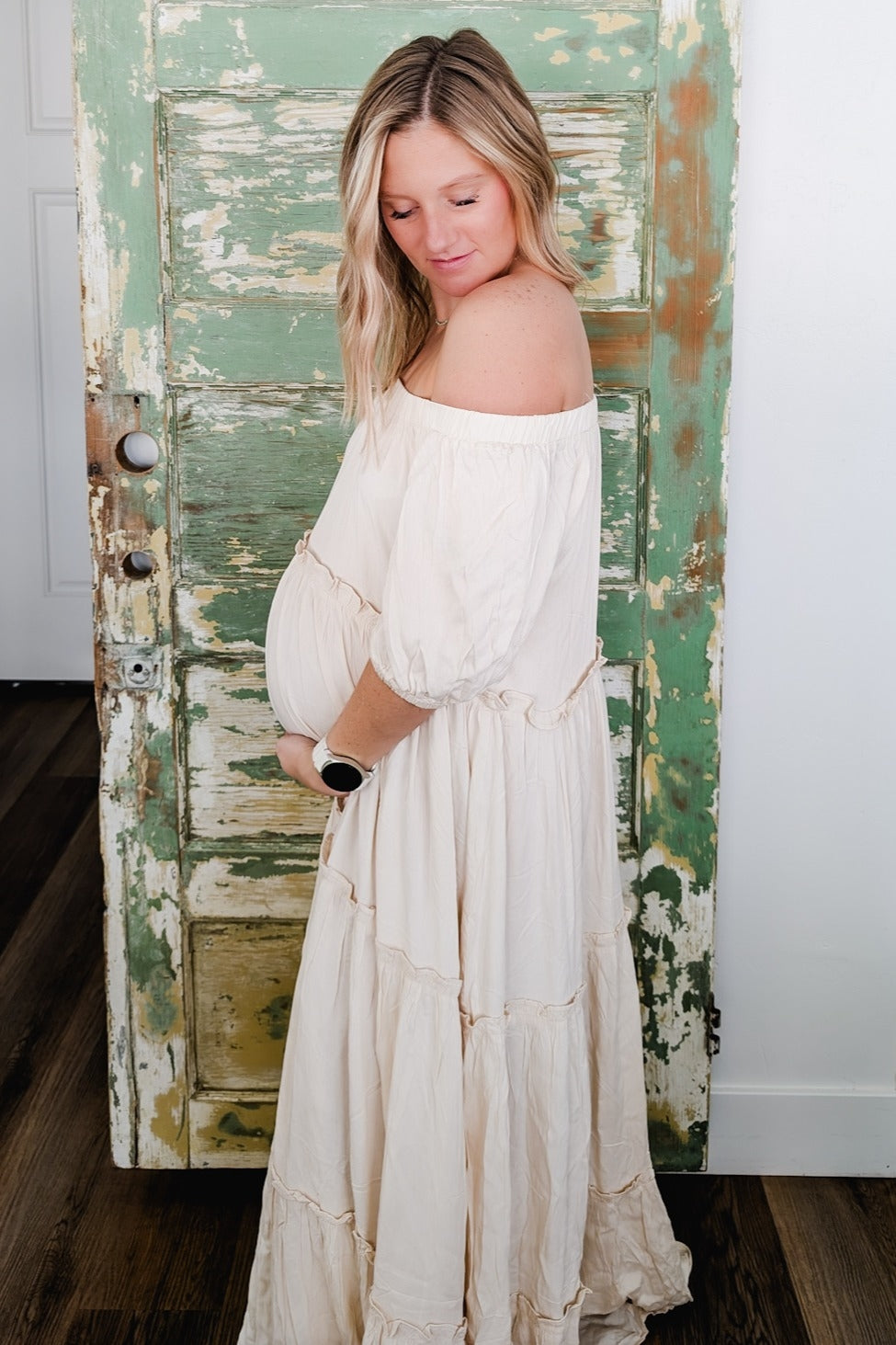 woman wearing a cream off-the-shoulder maternity dress with a flowing tiered design and holding her baby bump