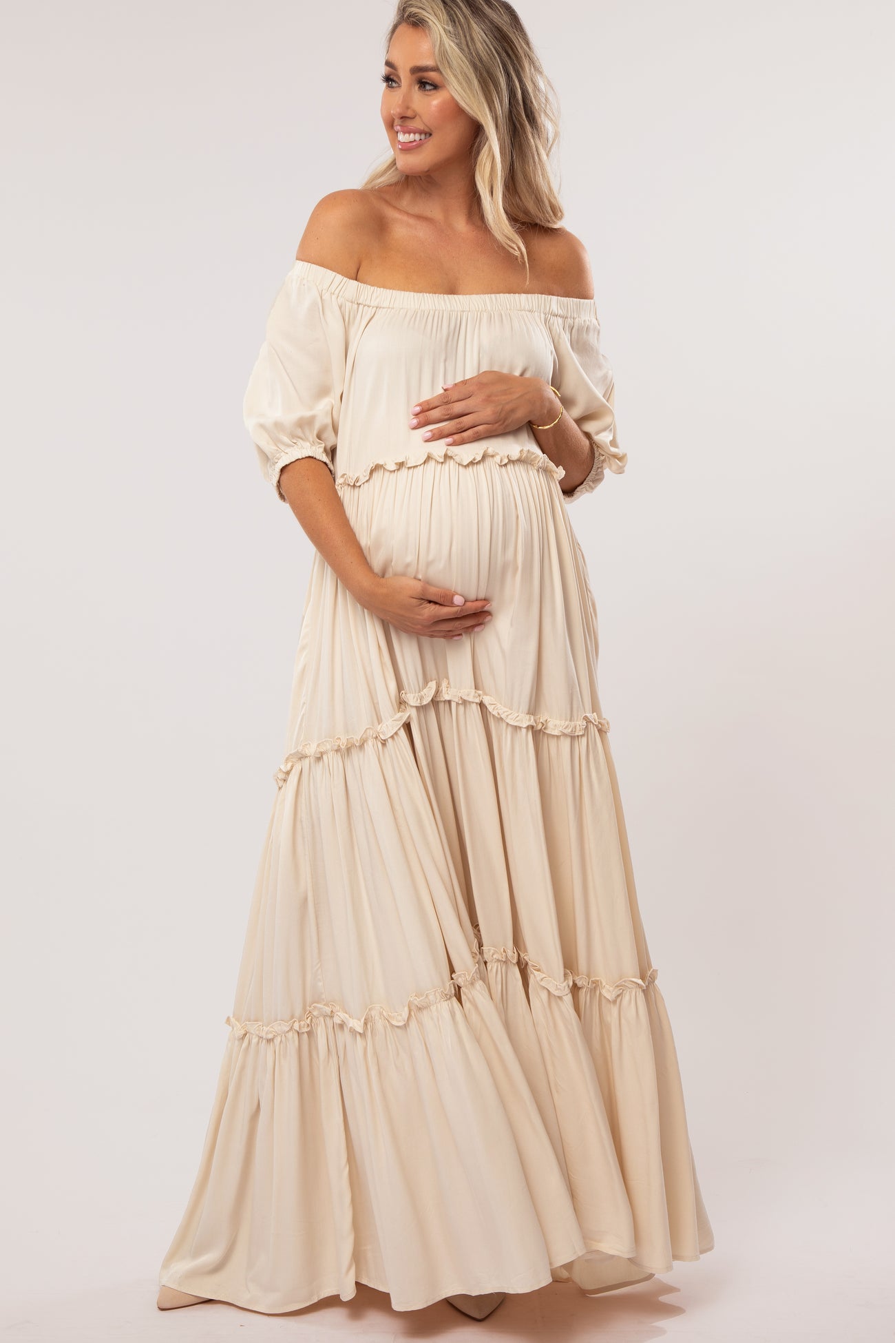 woman wearing a cream off-the-shoulder maternity maxi dress with ruffle details and cradling her baby bump