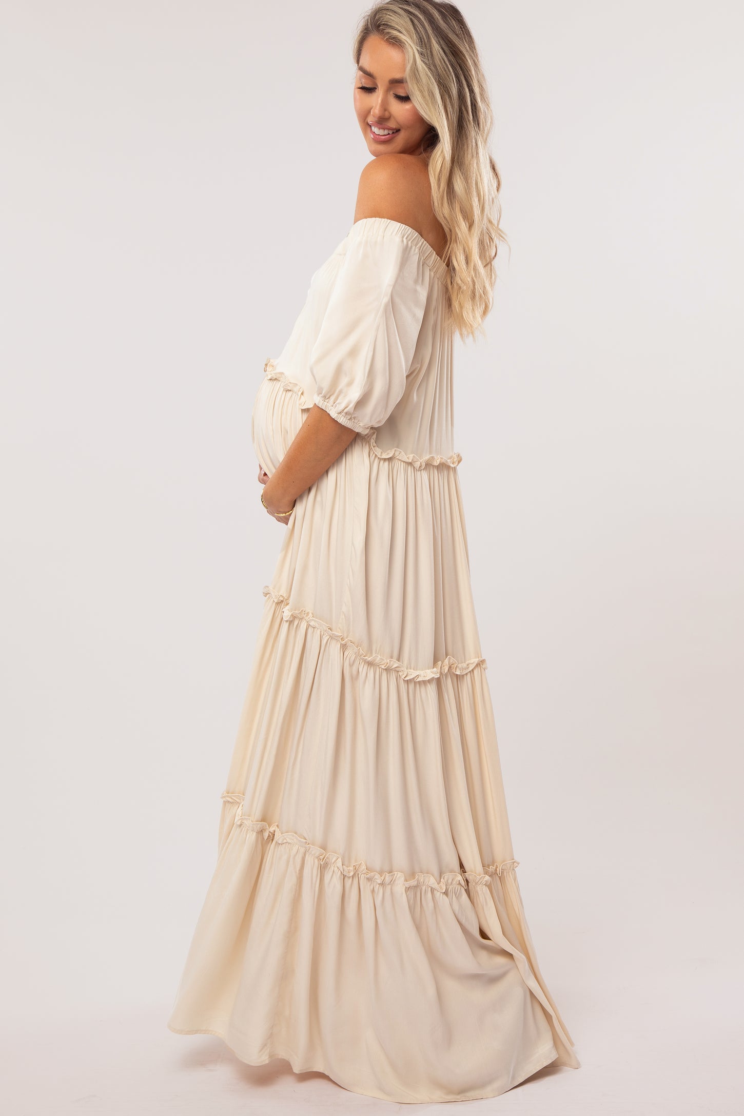 Pregnant woman in a cream off-the-shoulder tiered maternity maxi dress holding her baby bump