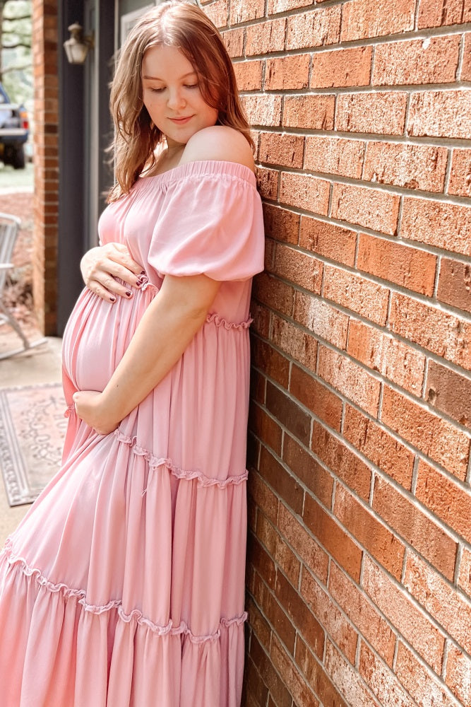 woman wearing a pink off-the-shoulder maternity dress with tiered ruffles while cradling her baby bump against a brick wall