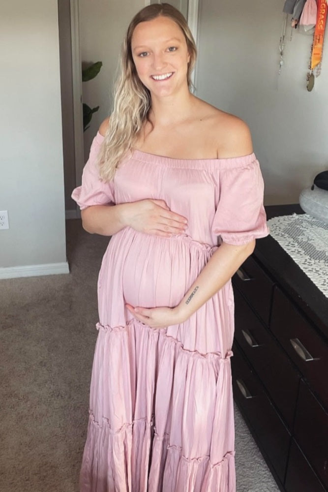 woman wearing a pink off-the-shoulder maxi dress and holding her baby bump, showcasing maternity wear