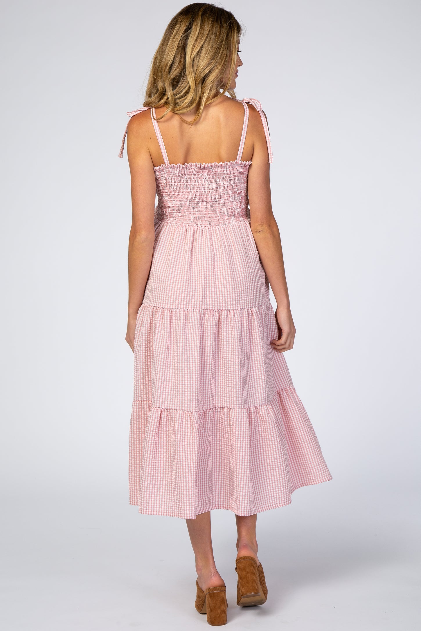 woman wearing a pink and white gingham maternity dress with tie shoulder straps and a tiered skirt, showing a baby bump
