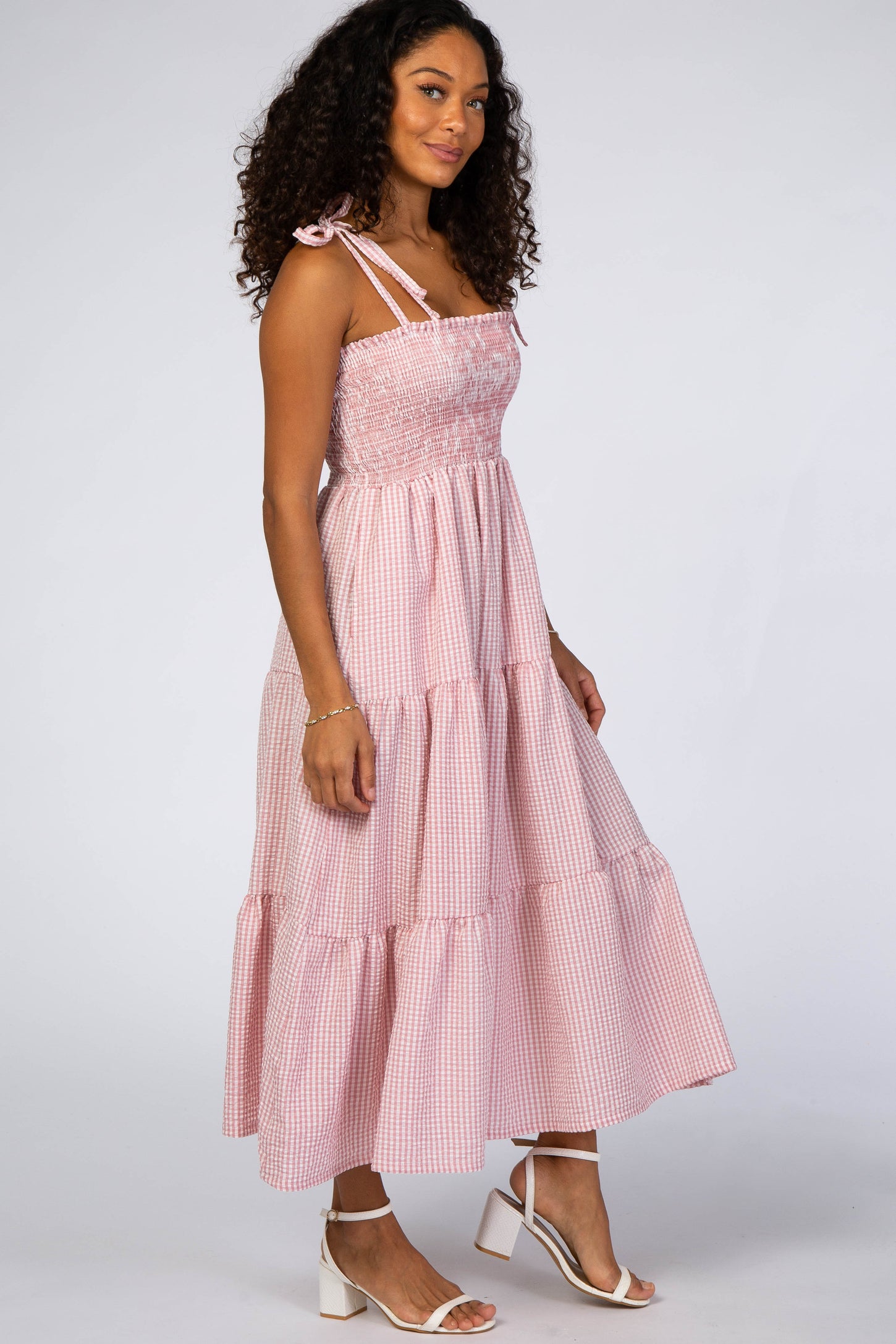 Woman wearing a pink gingham smocked tiered midi dress with tie straps and white block heel sandals