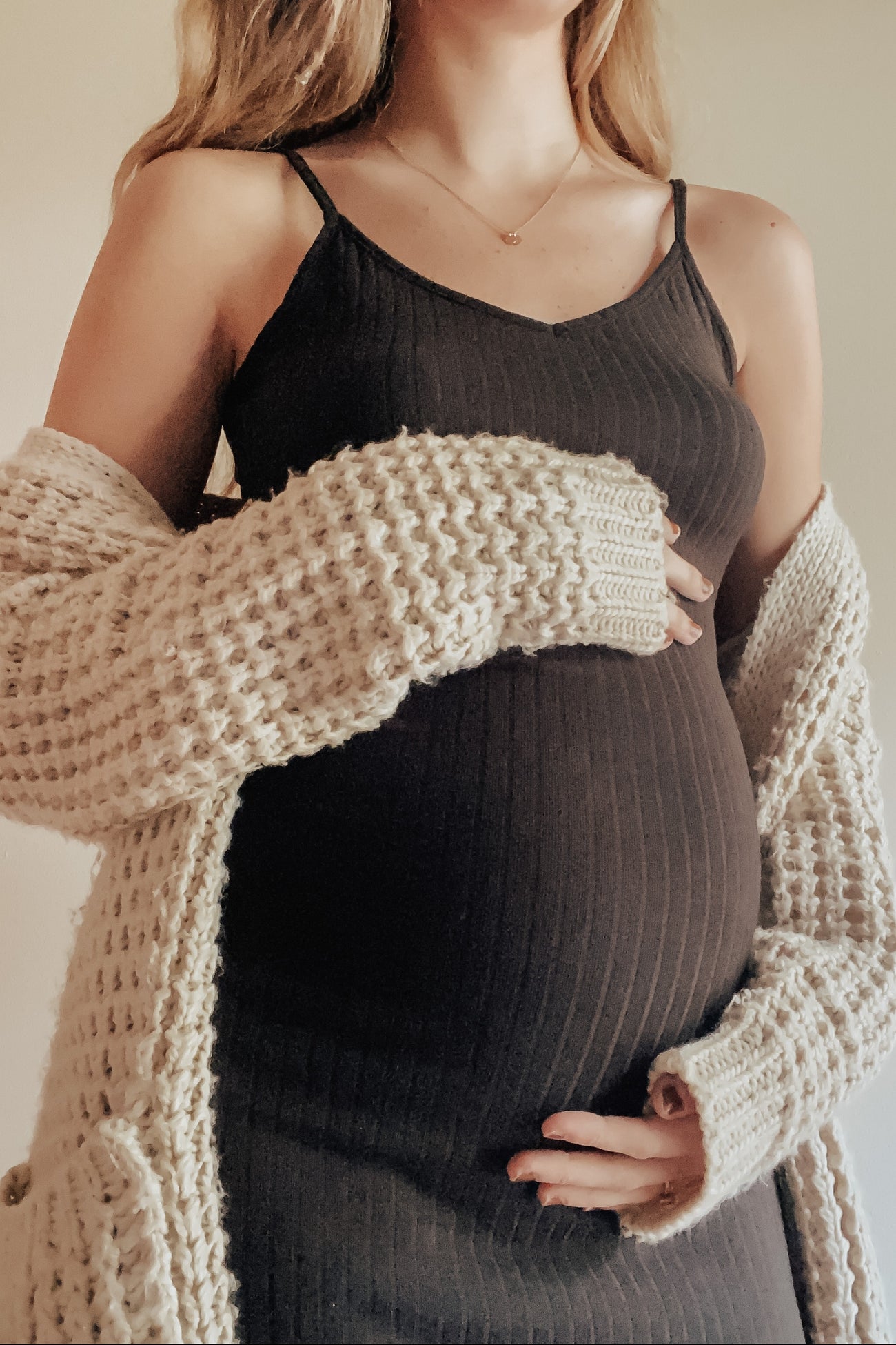 woman wearing a black ribbed maternity dress and a cream knitted cardigan, holding her baby bump