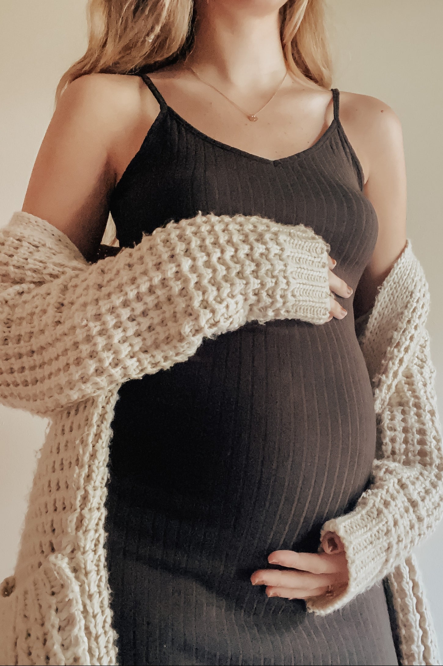 woman wearing a black ribbed maternity dress and a cream knitted cardigan, holding her baby bump