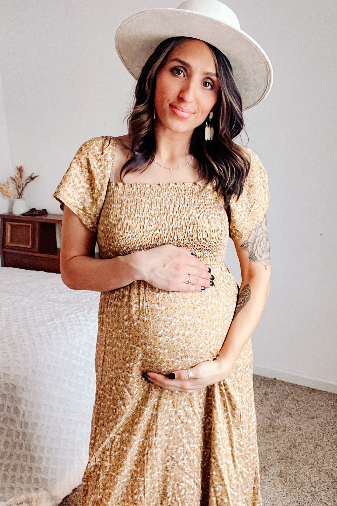 woman wearing a mustard yellow floral maternity dress and holding her baby bump while smiling