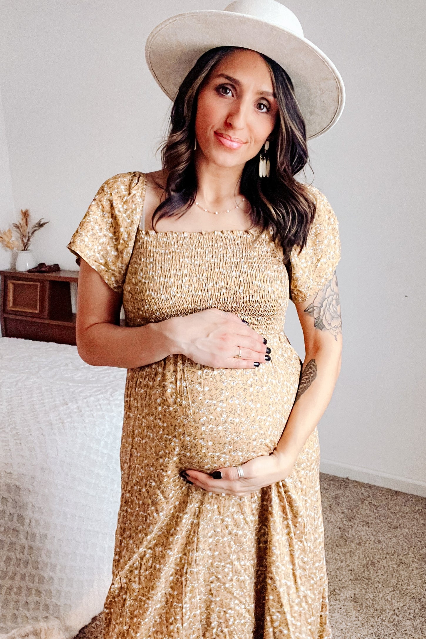woman wearing a mustard yellow floral maternity dress and holding her baby bump while smiling