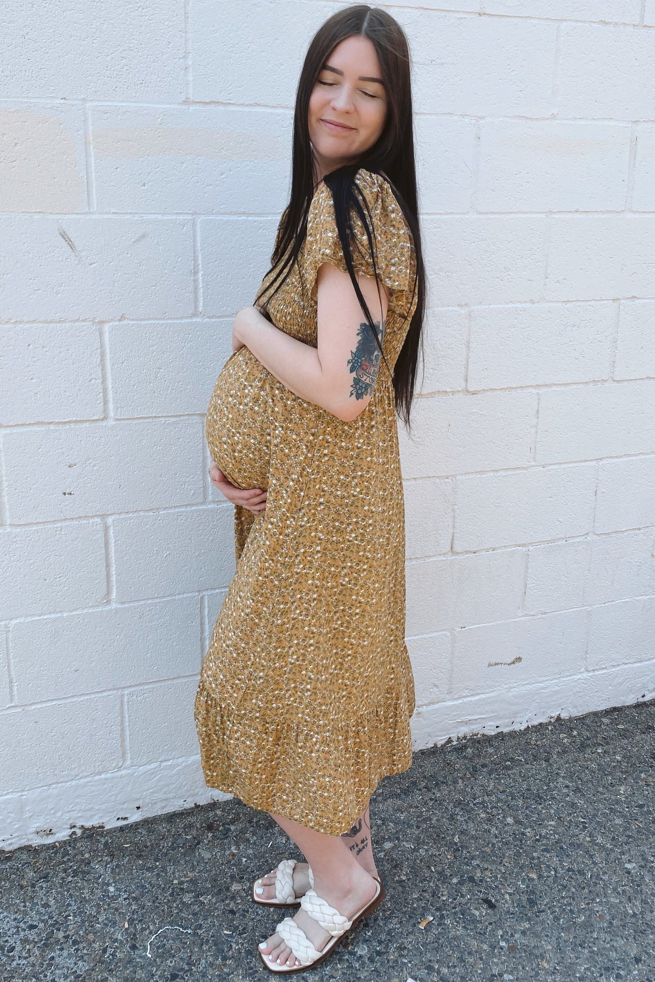woman wearing a mustard floral maternity dress, holding her baby bump, standing against a brick wall