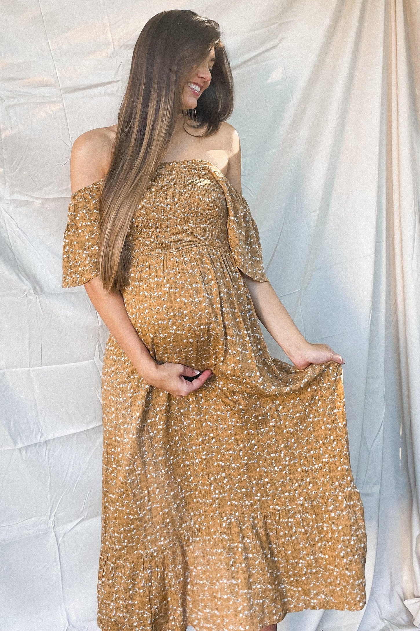 Pregnant woman in a mustard yellow floral off-the-shoulder maternity maxi dress holding her baby bump