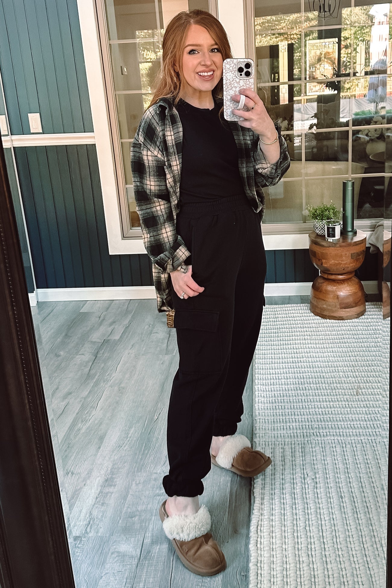 woman wearing a black t-shirt, plaid flannel shirt, and black cargo joggers with fluffy slippers, posing in a mirror