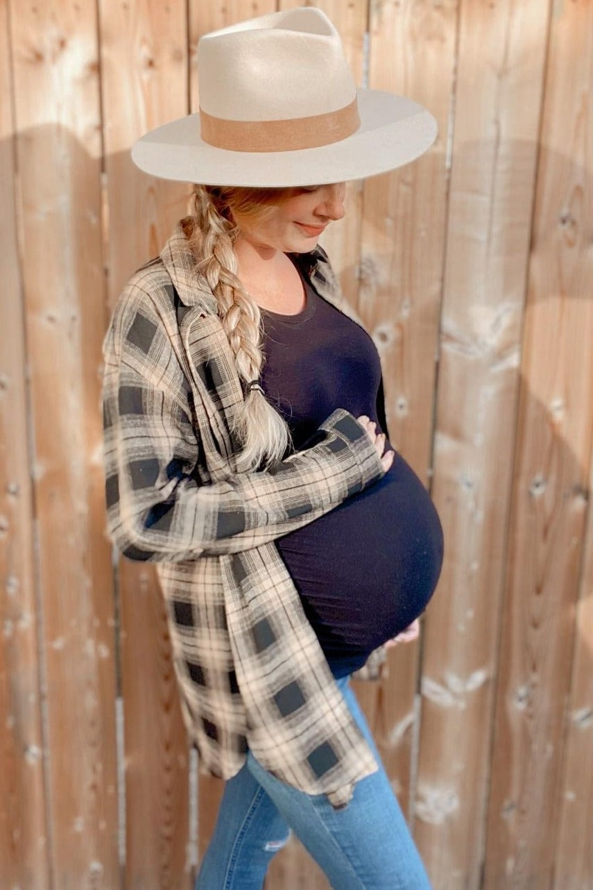 woman wearing a black maternity top with a plaid shirt, blue jeans, and a wide-brim hat, showcasing her baby bump