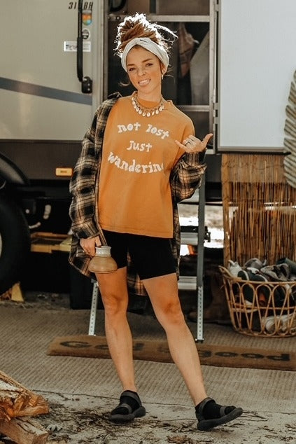 woman wearing an orange graphic t-shirt that says "not lost just wandering," black shorts, and a plaid shirt, posing outdoors