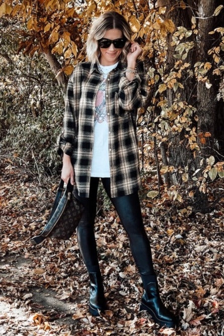 woman wearing a black and yellow plaid oversized shirt, graphic t-shirt, black leggings, and ankle boots, posing outdoors