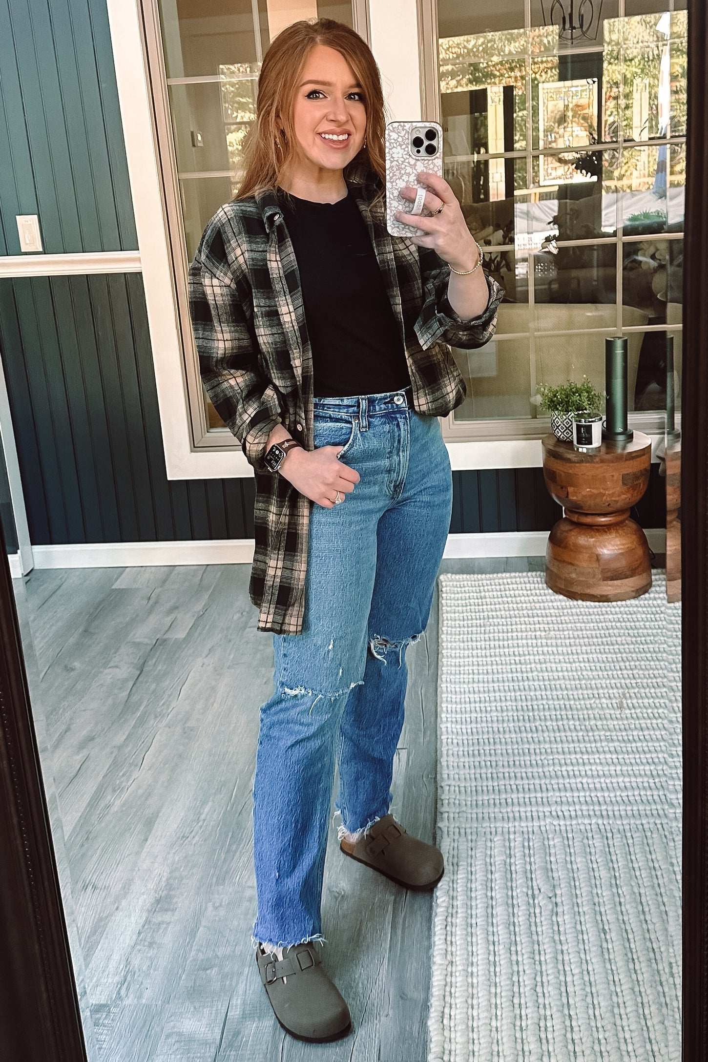 woman wearing a black t-shirt layered with a green plaid oversized shirt and distressed blue jeans, standing in front of a mirror