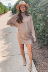 woman wearing a beige oversized sweater dress with long sleeves and a wide-brimmed hat, paired with tan ankle boots
