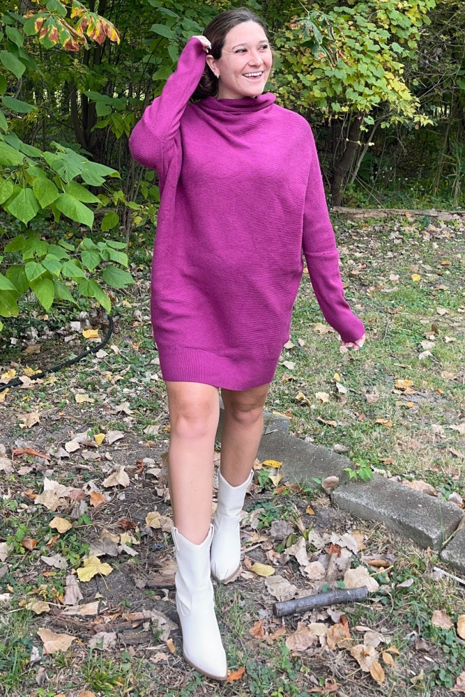woman wearing a bright pink oversized knit sweater dress and white ankle boots, walking through a leafy outdoor area