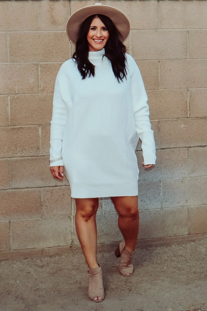 woman wearing a white long sleeve sweater dress, tan wide brim hat, and suede ankle boots against a brick wall