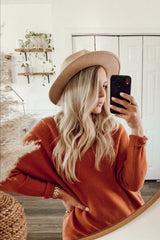 woman with long blonde hair wearing a tan wide-brim hat and orange sweater taking a mirror selfie