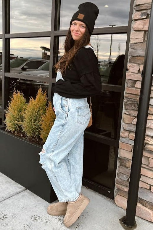 woman wearing a black long sleeve top and distressed light blue maternity overalls, showing her baby bump, standing outside
