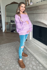 pregnant woman in purple off-the-shoulder sweater and ripped jeans standing by a fireplace