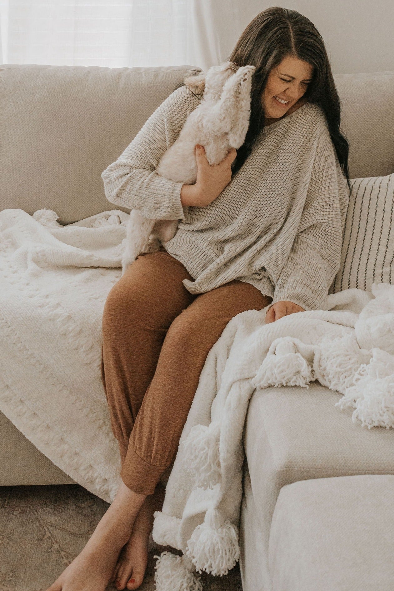 woman in a cozy beige oversized sweater and brown lounge pants sitting on a couch and holding a small white dog