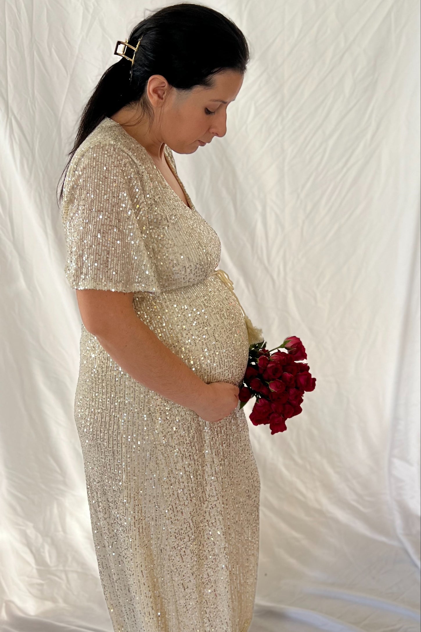 woman wearing a sparkling gold maternity gown and holding a bouquet of red roses, showcasing her baby bump