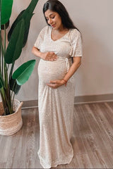woman wearing a shimmering beige maternity gown and holding her baby bump while standing next to a plant