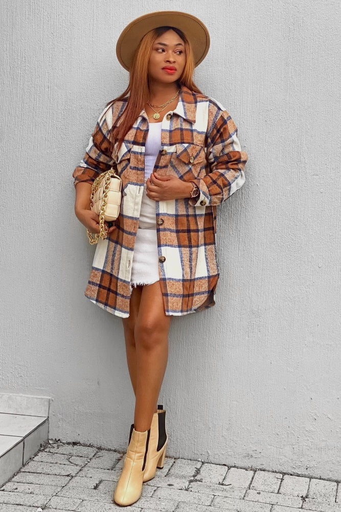 Woman in brown plaid shacket, white mini skirt, and tan fedora hat posing against a grey wall