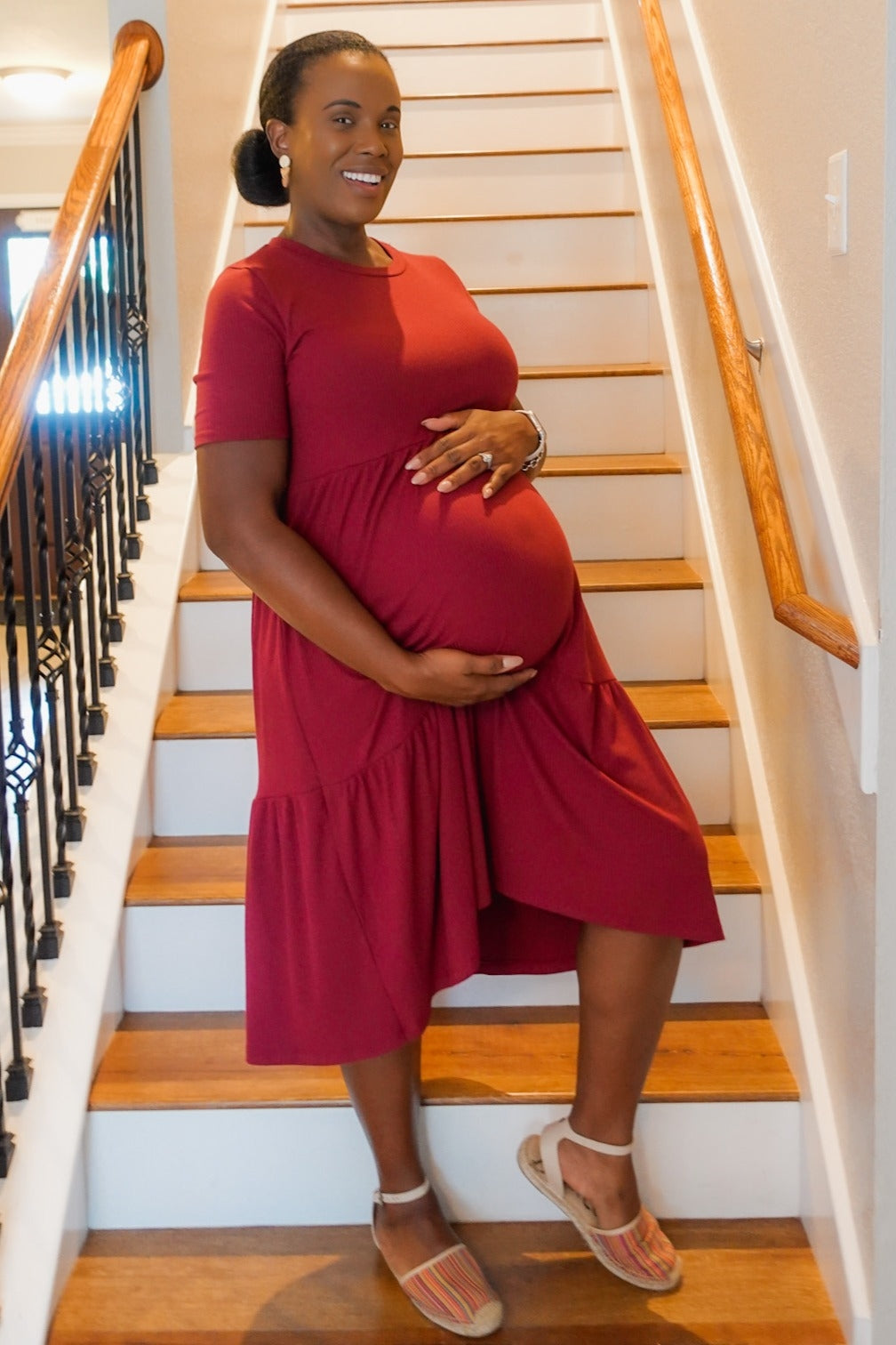 Smiling pregnant woman in a red maternity dress posing on a wooden staircase while holding her baby bump