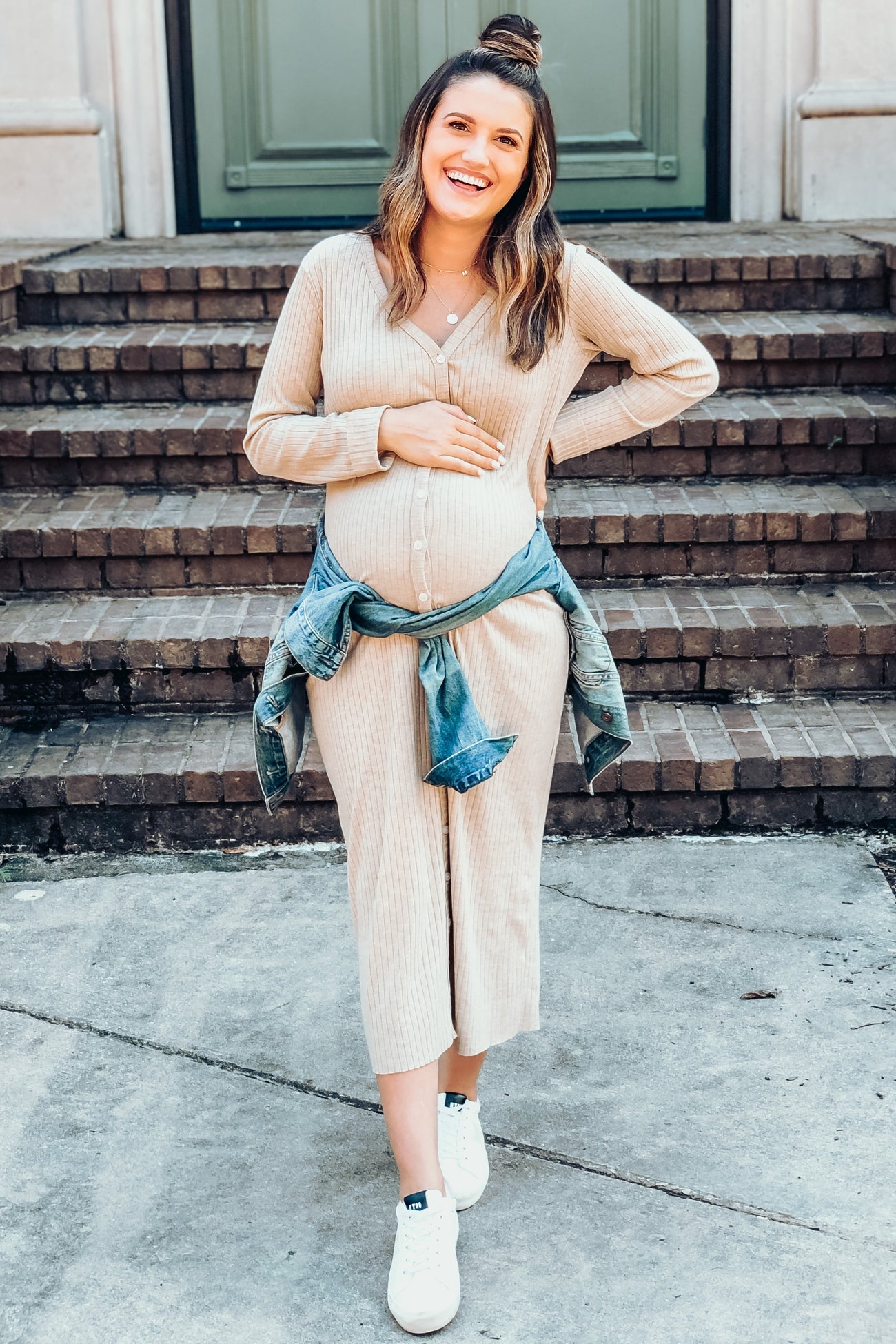 smiling pregnant woman in beige ribbed maternity dress and denim jacket standing on stone steps