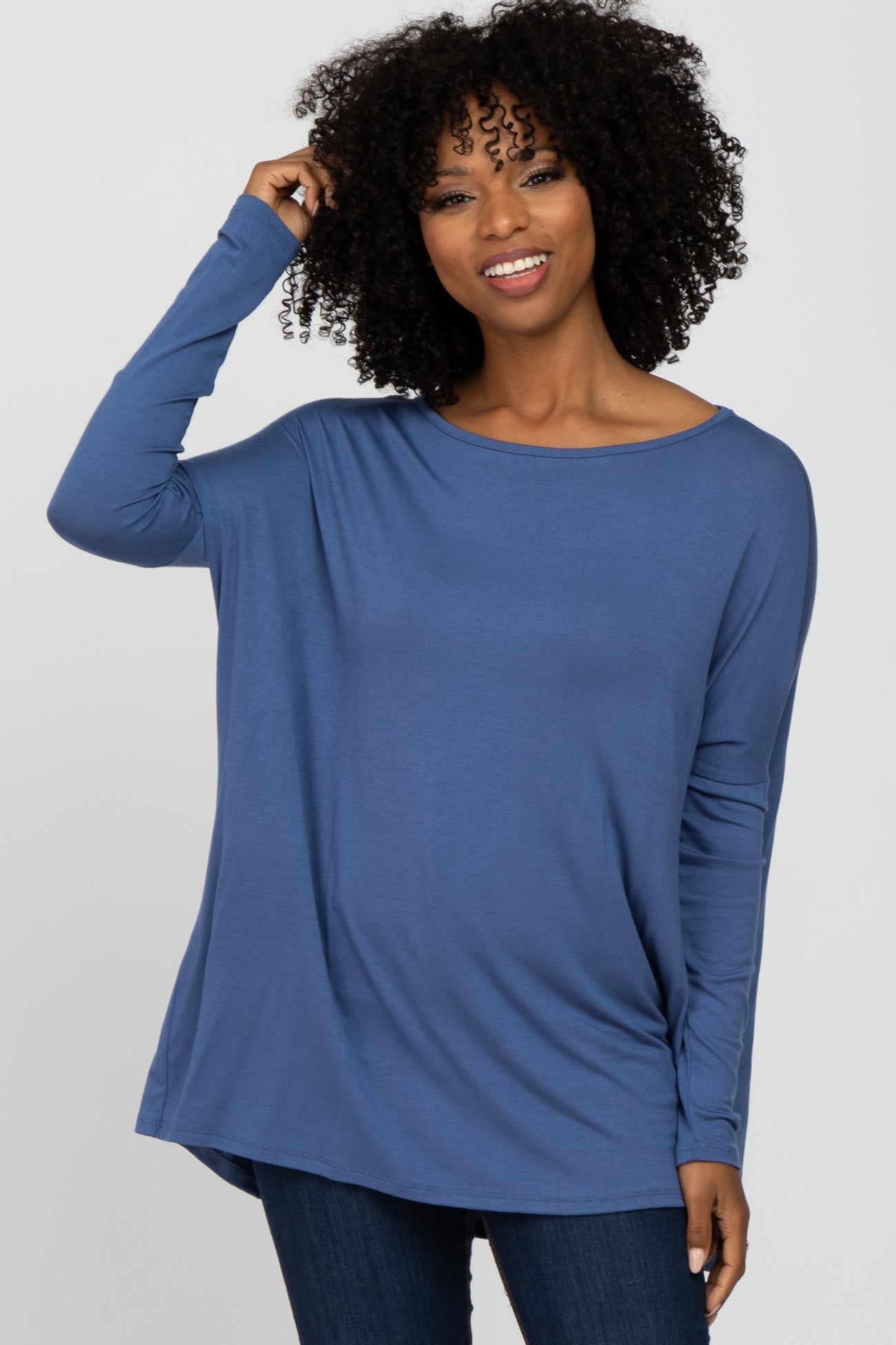 Woman with curly hair wearing a long-sleeved blue tunic top and dark jeans against a white background