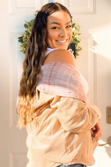 smiling woman with long wavy hair wearing a tan corduroy jacket and pink plaid shirt standing by a door