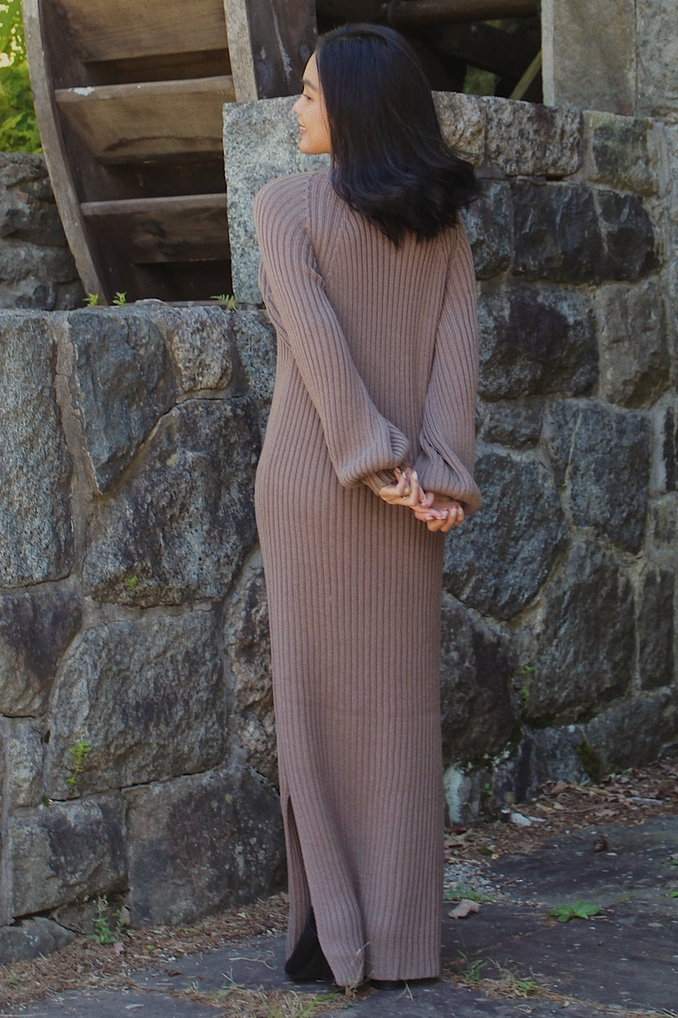 woman wearing a long brown ribbed knit sweater dress with side slits, standing outdoors with hands behind her back