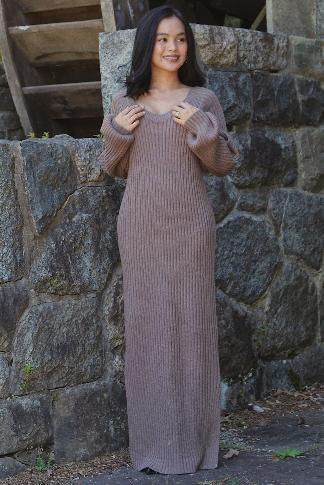 woman wearing a taupe ribbed long sleeve maxi dress, standing against a stone wall