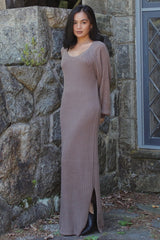 woman wearing a taupe ribbed maxi dress with long sleeves and side slits, standing against a stone wall