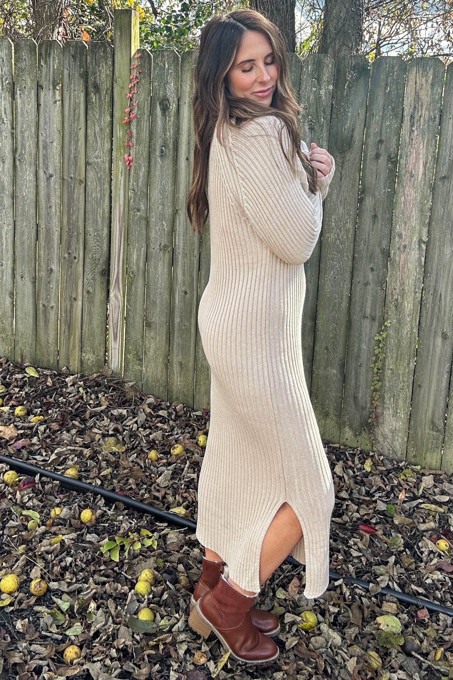 woman wearing a beige ribbed sweater dress with side slits and brown ankle boots, standing outdoors among fallen leaves