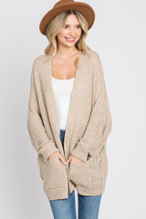 woman wearing a beige oversized knit cardigan with a white tank top and blue jeans, smiling and wearing a brown hat