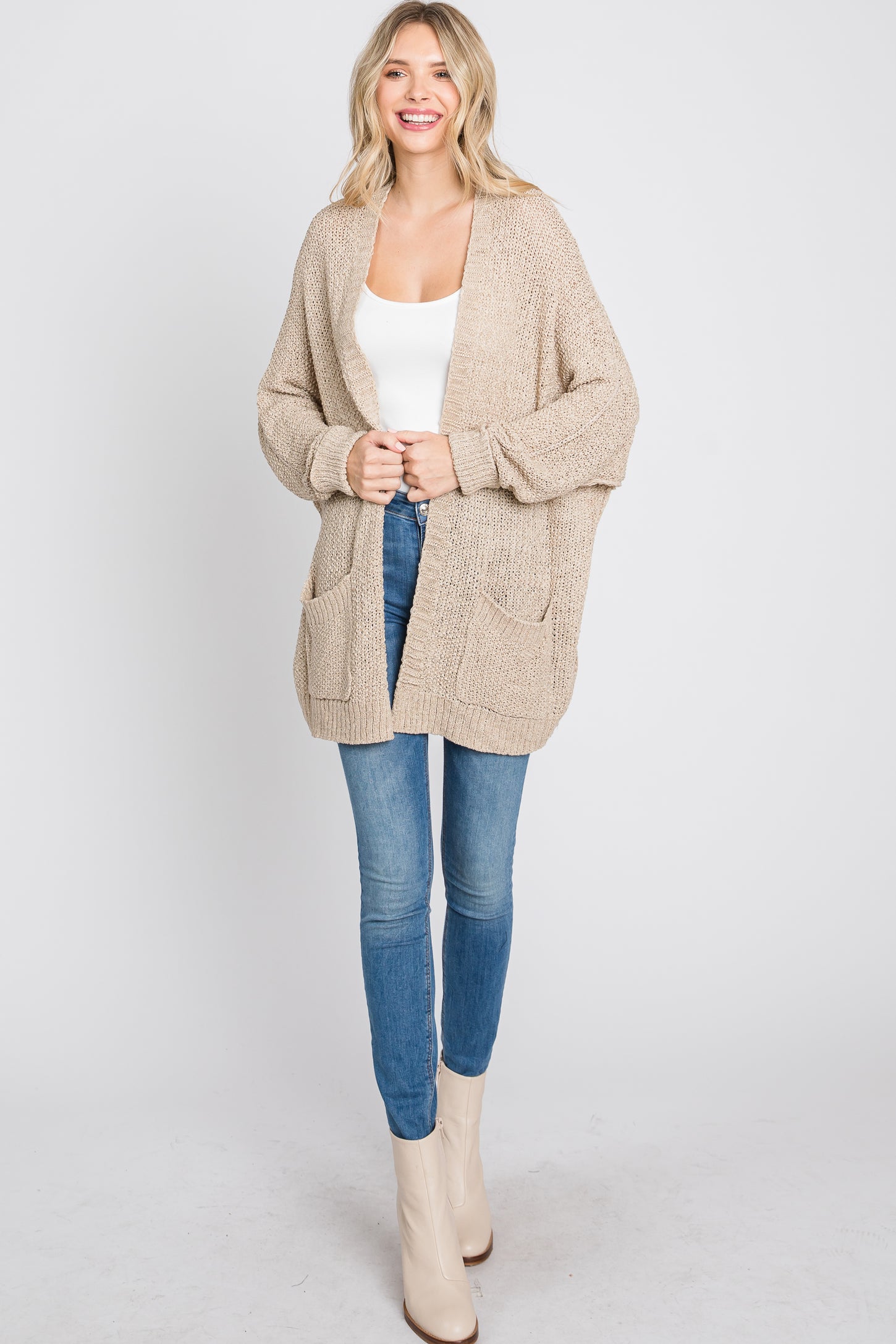 woman wearing a knitted beige cardigan with pockets, paired with a white tank top and blue skinny jeans