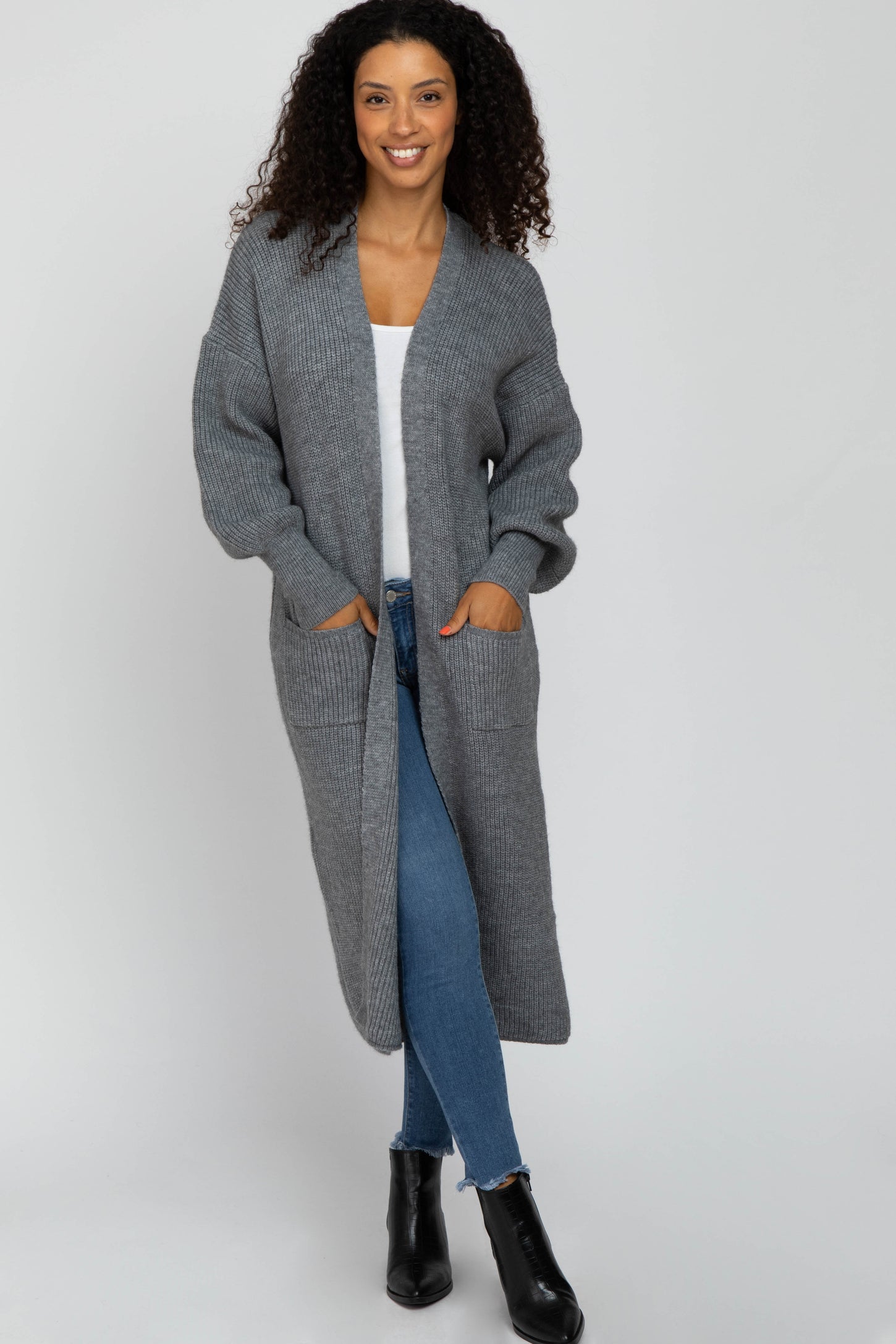 woman wearing a long gray knit cardigan with pockets, white top, and blue jeans, standing and smiling