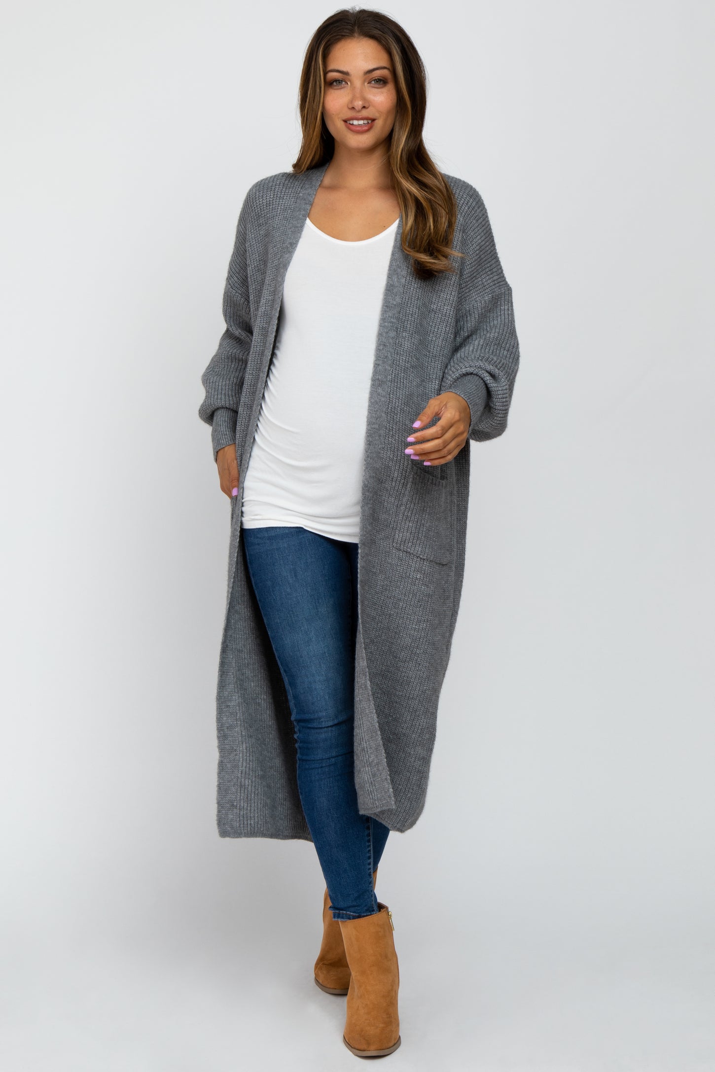 woman wearing a long gray knit maternity cardigan over a white tank top and blue jeans, showing off her baby bump