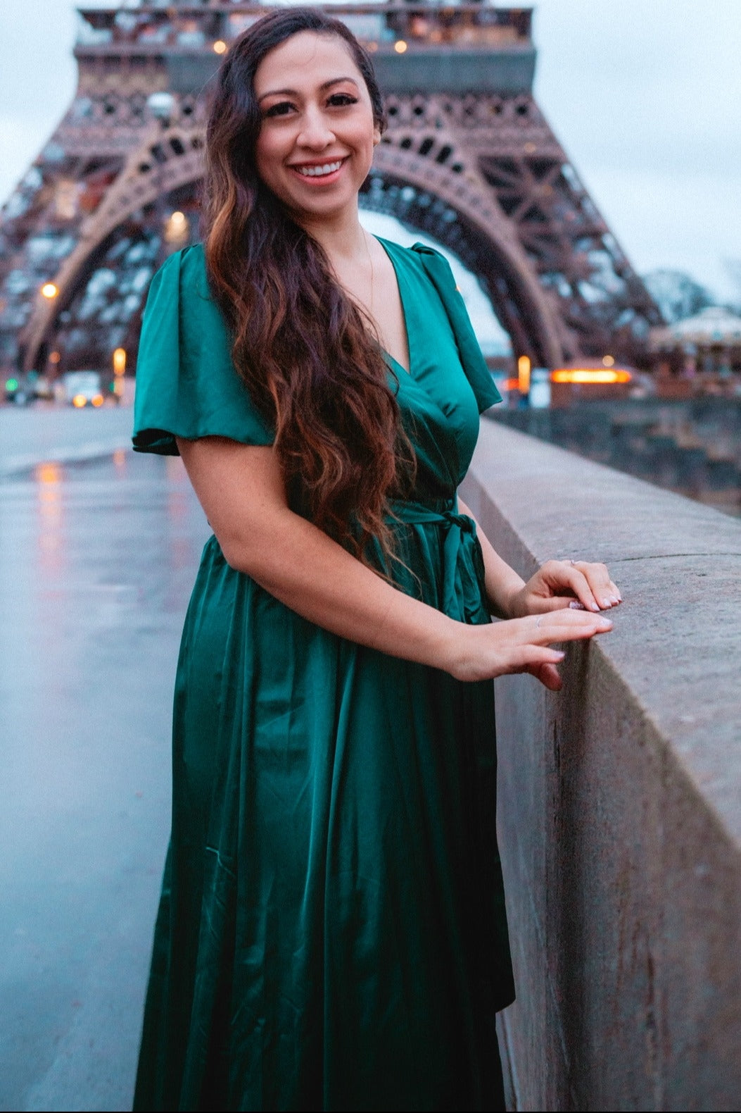 woman wearing a green satin wrap dress, standing in front of the eiffel tower, smiling with long wavy hair